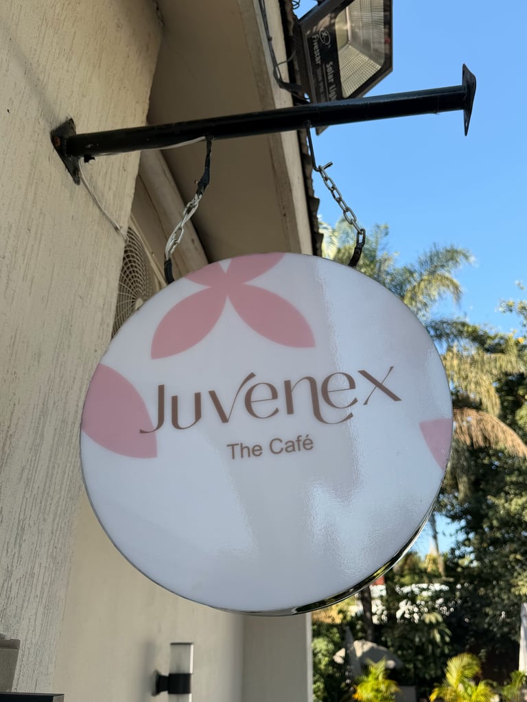 Round white and pink Juvenex the Café hanging sign on a building exterior with clear blue sky and trees